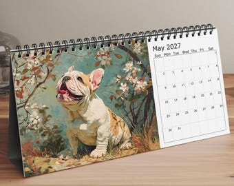 May include: A spiral-bound desk calendar featuring a painting of a cream and white French Bulldog sitting among flowering branches. The calendar page displays the month of May 2027 with a grid of dates from the 1st to the 31st in black text.