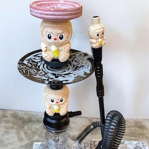 May include: A decorative hookah with a clear glass base and black stem. The hookah features cartoon character figurines holding yellow items. The top tray is pink, and the hose is gray and black.