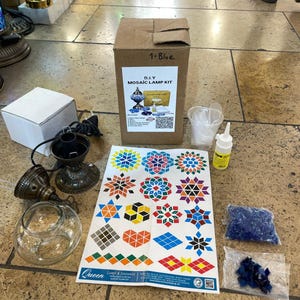 Op de afbeelding: Een doe-het-zelf mozaïek lamp kit met een bruine kartonnen doos met het label "1-Blauw". De kit bevat een lampvoet, glasstukken, lijm en een vel kleurrijke mozaïekontwerpen. De ontwerpen bevatten geometrische vormen en bloempatronen. De kit is voor het maken van een mozaïeklamp.