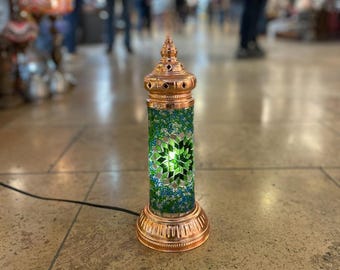 May include: A decorative table lamp with a copper-colored base and top. The cylindrical body is covered in a mosaic of green, blue, and clear glass. The lamp is lit, showcasing a star-shaped design in the center. A black power cord extends from the base.