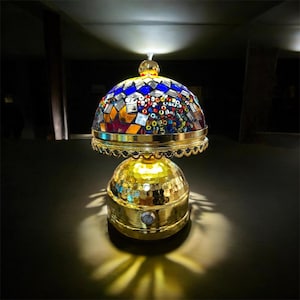Op de afbeelding: Een decoratieve tafellamp met een mozaïek glazen kap in tinten blauw, rood, oranje en helder. De voet en de afwerking zijn goudkleurig. De lamp is verlicht en geeft een warme gloed.