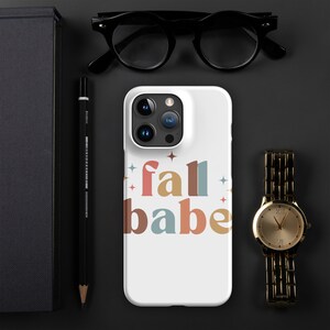 Puede incluir: Funda de teléfono blanca con el texto "fall babe" en una fuente retro con acentos de estrellas. Un reloj de pulsera dorado y gafas negras están al lado de la funda del teléfono. Un lápiz negro y un cuaderno también están en la imagen.