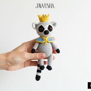 May include: A crocheted lemur wearing a blue and yellow cape and a gold crown. The lemur is grey with black eyes and a black and white striped tail.