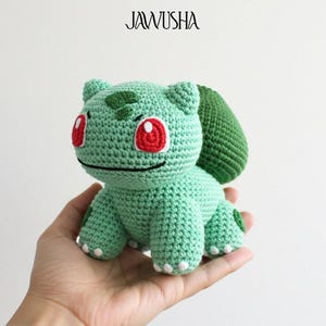 Puede incluir: Un peluche de Bulbasaur tejido a mano en tonos verdes, con ojos rojos y una expresión sonriente. El juguete hecho a mano tiene un bulbo en la espalda y se sostiene en una mano. La imagen incluye el texto "JAWUSHA" y "DIGITAL PDF PATTERN".