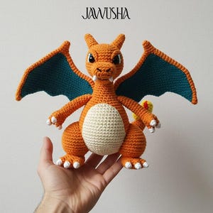 May include: Crocheted orange dragon with teal wings and a cream-colored belly. The dragon is held in a hand. The word "JAWUSHA" is above the dragon. The image also includes the text "DIGITAL PDF PATTERN".
