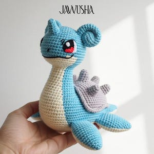 Puede incluir: Peluche de Lapras tejido a ganchillo en tonos azul, crema y gris. El juguete hecho a mano presenta detalles como ojos grandes, caparazón y aletas. El texto "JAWUSHA" está en la parte superior y un logotipo de "DIGITAL PDF PATTERN" en la parte inferior.