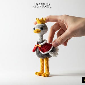 May include: A crocheted gray ostrich wearing a red and white cape and a yellow crown. The ostrich is standing on a white surface.