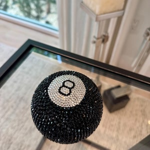May include: A black and silver decorative eight ball, covered in sparkling crystals. The number eight is displayed in a white circle. The ball is resting on a glass surface.
