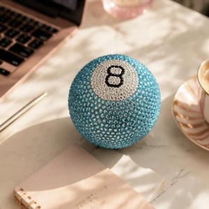May include: A spherical eight ball covered in light blue rhinestones with a white circular patch on the front featuring a black number eight. The surface of the ball is entirely encrusted with small, reflective crystals that catch the light.