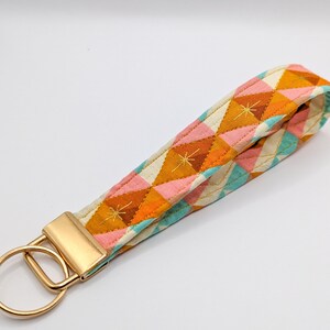 May include: A gold key chain with a fabric strap featuring a geometric pattern in shades of orange, pink, and blue. The fabric has a quilted texture and a subtle star design.