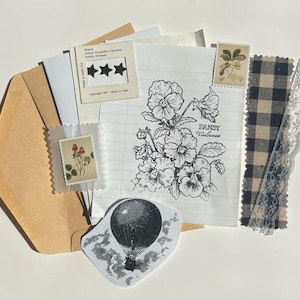 May include: A selection of vintage ephemera including stamps, patterned fabric, and a botanical illustration. A brown envelope holds various paper scraps, a black and white hot air balloon sticker, and a lined paper with a floral drawing.