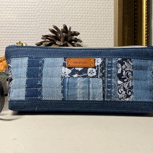 Pode incluir: Uma bolsa com zíper de patchwork de ganga com uma etiqueta de couro castanho que diz "Handmade". A bolsa apresenta uma variedade de tecidos de ganga azul em diferentes padrões e tons. Um fecho de correr dourado e uma pinha são visíveis.