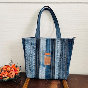 Op de afbeelding: Een denim tote bag met verticale strepen in verschillende tinten blauw. De tas heeft twee handvatten en een klein leren label met het woord "handmade" en het jaar "1969". De tas staat op een houten oppervlak.