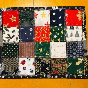 Puede incluir: Un posavasos cuadrado de patchwork con un marco negro, hecho con una variedad de telas con temática navideña. Las telas incluyen rojo, verde, blanco y negro con varios patrones que incluyen estrellas, copos de nieve, árboles y acebo.