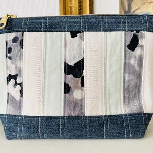 Peut inclure: Une trousse de maquillage rectangulaire avec un motif patchwork. Le sac présente des rayures verticales dans les tons de blanc, gris et noir, avec des accents en jean en haut et en bas. Une fermeture éclair dorée sécurise le dessus.