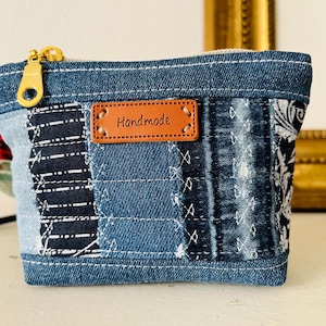 Può includere: Pochette in denim fatta a mano con cerniera dorata. La pochette presenta un design patchwork con vari modelli in denim e un'etichetta in pelle marrone con la scritta "Handmade". La combinazione di colori è blu con accenti neri e bianchi.