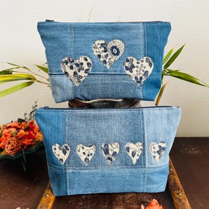 Op de afbeelding: Twee denim ritszakjes met hartapplicaties. De zakjes zijn gemaakt van verschillende tinten blauw denim. Elk zakje heeft witte harten met een blauw bloemmotief. De zakjes zijn op elkaar gestapeld.
