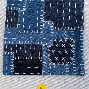 May include: A square patchwork coaster made of blue denim fabric with white stitching. The coaster is made of different pieces of denim sewn together in a patchwork pattern. The stitching is done in a variety of patterns, including straight lines, crosses, and Xs. The coaster is a light blue colour and has a slightly textured surface.