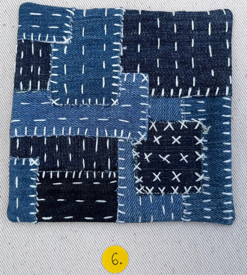 May include: A square patchwork coaster made of blue denim fabric with white stitching. The coaster is made of different pieces of denim sewn together in a patchwork pattern. The stitching is done in a variety of patterns, including straight lines, crosses, and Xs. The coaster is a light blue colour and has a slightly textured surface.