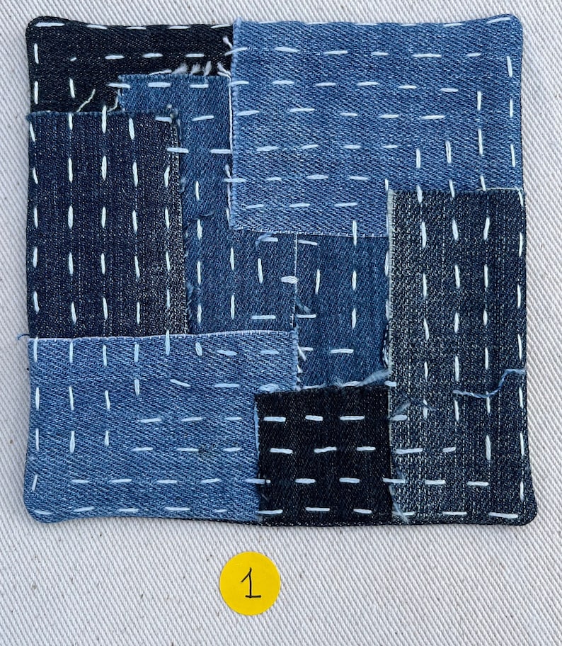 May include: A square patchwork cushion made from blue denim fabric with white stitching. The cushion is made up of different sized squares of denim, some of which are darker than others. The cushion is sitting on a white surface with the number "1" written on a yellow circle.