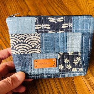 Può includere: Una piccola pochette in denim patchwork con una chiusura a zip. La pochette è realizzata in diverse tonalità di denim blu e presenta un'etichetta in pelle marrone che dice "Handmade".
