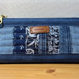 Op de afbeelding: Een denim patchwork etui met een gouden rits. De etui heeft verschillende blauwe denim patronen, waaronder anker- en bloemmotieven. Een klein bruin leren label leest "Handmade".