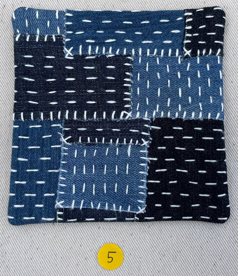 May include: A blue and black patchwork fabric square with white stitching. The fabric is made of denim and has a variety of textures. The square is labelled with the number "5".