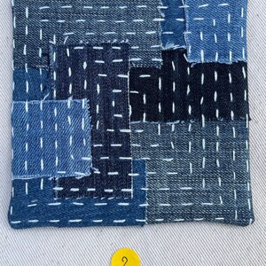 May include: A blue and black patchwork fabric square with white stitching. The fabric is made of denim and has a textured surface. The square is numbered "2" in a yellow circle.