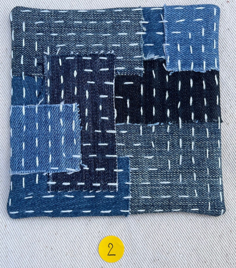 May include: A blue and black patchwork fabric square with white stitching. The fabric is made of denim and has a textured surface. The square is numbered "2" in a yellow circle.