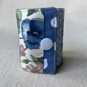 Peut inclure: Une petite pochette en tissu bleu et blanc avec un imprimé floral et une fermeture à bouton blanc. La pochette est composée de deux couches de tissu, l'une avec un imprimé floral et l'autre avec des pois blancs sur fond bleu.