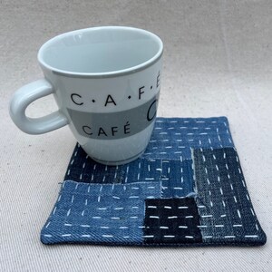 May include: A white ceramic mug with black text that reads "C.A.F.E. CAFE" sits on a blue and white denim coaster. The coaster is made of patchwork denim squares with white stitching.