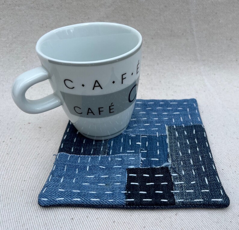 May include: A white ceramic mug with black text that reads "C.A.F.E. CAFE" sits on a blue and white denim coaster. The coaster is made of patchwork denim squares with white stitching.