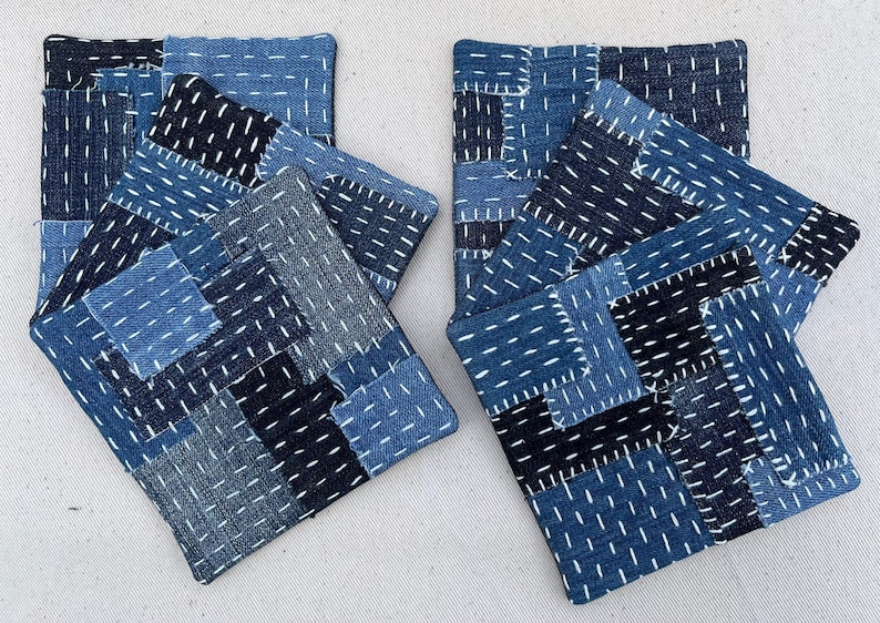 May include: Set of six blue and black denim coasters with white stitching. The coasters are made of patchwork fabric and are arranged in two stacks of three.