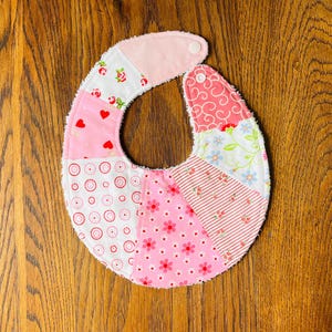 Peut inclure: Une bavette de bébé en patchwork rose et blanc avec une bordure en tissu éponge blanc. La bavette présente une variété de motifs floraux et géométriques.