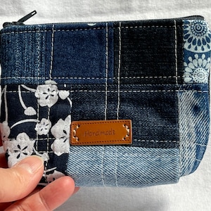 Peut inclure: Une petite pochette zippée en jean patchwork avec une étiquette en cuir marron qui dit "Handmade". La pochette est faite de différentes nuances de jean bleu et a un motif floral blanc sur un côté.