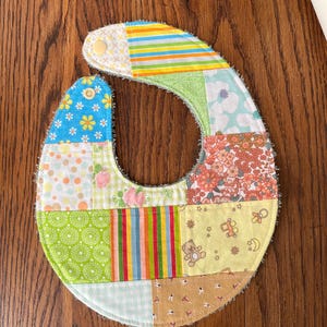 Op de afbeelding: Een patchwork baby-slabbetje met verschillende kleurrijke patronen, waaronder bloemen, geometrische en gestreepte ontwerpen. Het slabbetje is gemaakt van zacht, absorberend materiaal en heeft een drukknoop sluiting.