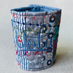 Peut inclure: Un bracelet en tissu bleu et blanc avec le nom "Jola" brodé en fil bleu.