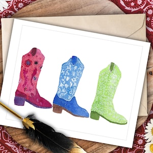 Peut inclure: Illustration à l'aquarelle de trois bottes de cow-boy rouges, bleues et vertes. Les bottes sont décorées de motifs floraux et cachemire. L'image est sur une carte blanche avec une enveloppe marron, reposant sur une surface en bois avec un bandana rouge.