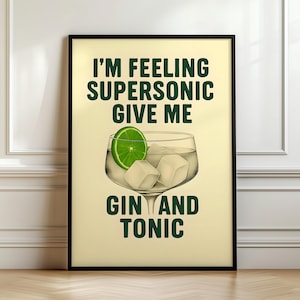 Op de afbeelding: Een ingelijste poster met de tekst "I'M FEELING SUPERSONIC GIVE ME GIN AND TONIC" in het groen. De afbeelding toont een gin-tonic cocktail met ijsblokjes en een schijfje limoen. De poster zit in een zwarte lijst.