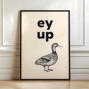 Könnte beinhalten: Gerahmter Kunstdruck mit dem Schriftzug "ey up" über einer Entenillustration. Text und Ente sind schwarz auf cremefarbenem Hintergrund. Der Druck befindet sich in einem schwarzen Rahmen und wird auf einem Holzboden vor einer weißen Wand ausgestellt.
