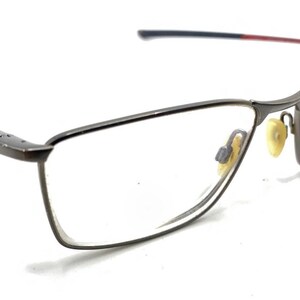 May include: Rectangular eyeglasses with a dark metallic frame, clear lenses, and adjustable nose pads. The temples feature a thin red accent near the hinges, and the frame arms transition to a dark blue color at the tips, providing a sleek look.
