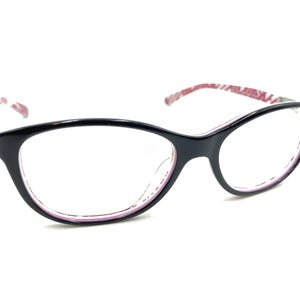 Puede incluir: Gafas negras con forma de ojo de gato. Las monturas son negras con interior rosa y patillas estampadas. Las lentes son transparentes. Las gafas están sobre un fondo blanco.