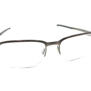 Puede incluir: Un par de gafas con montura de metal gris oscuro y lentes transparentes. Las gafas tienen forma rectangular con un diseño moderno. Las patillas son finas y se extienden hasta los extremos.