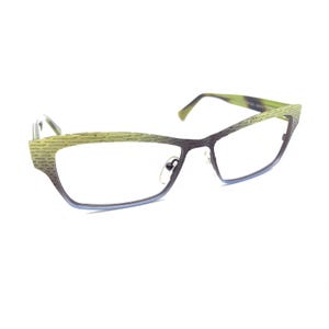 Puede incluir: Gafas con una montura degradada que pasa de un verde lima texturizado en la parte superior a un morado oscuro sólido en la inferior. La montura tiene una forma de ojo de gato con lentes transparentes, almohadillas integradas y varillas finas.