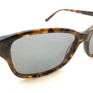 May include: Tortoiseshell-patterned sunglasses with gray lenses. The frames have a classic cat-eye shape with a dark brown and gold color scheme. The temples are dark brown with gold accents.