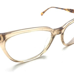 May include: Eyeglasses with a translucent beige frame and a cat-eye shape. The temples feature a brown and black tortoiseshell pattern. The clear lenses are set within the plastic frame, which connects to thin arms that extend outward to the sides.