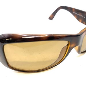 May include: Tortoiseshell sunglasses featuring a thick, rounded frame with a glossy finish and amber-tinted lenses. The temples display a gold-toned metallic emblem on the side, and the inner arm includes printed model information in white text.