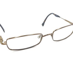 Op de afbeelding: Een bril met een goudkleurig metalen montuur. De glazen zijn rechthoekig met afgeronde randen. De veren hebben een donkerbruin en beige patroon. De bril staat tegen een witte achtergrond.