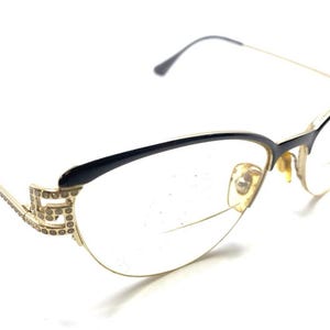 May include: Cat-eye glasses featuring a black upper frame and a gold-tone lower rim. The temples are gold-tone with a decorative Greek key pattern embellished with small clear crystals. The nose pads are gold-tone and the lenses are clear.