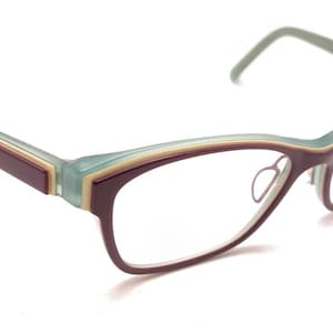 Puede incluir: Gafas con forma de ojo de gato. Las monturas son de color burdeos oscuro con un acento verde claro y beige en la parte superior y las varillas. Las lentes son transparentes y las varillas son burdeos.